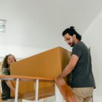 From below of couple in casual clothes carrying big carton box together while moving packed personal items into new apartment