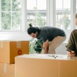 Positive young woman and focused man carrying cardboard boxes in new apartment on moving day
