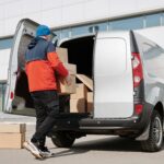 Man in colorful jacket loading cardboard boxes into a van outside an office building.