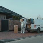 A courier in protective gear delivers packages from a van to a residential house.