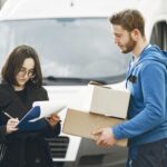 A courier in blue hands packages to a woman signing documents in front of a delivery van.