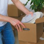 Crop anonymous male in casual clothes packing fragile plates in carton box before moving into new flat in daylight